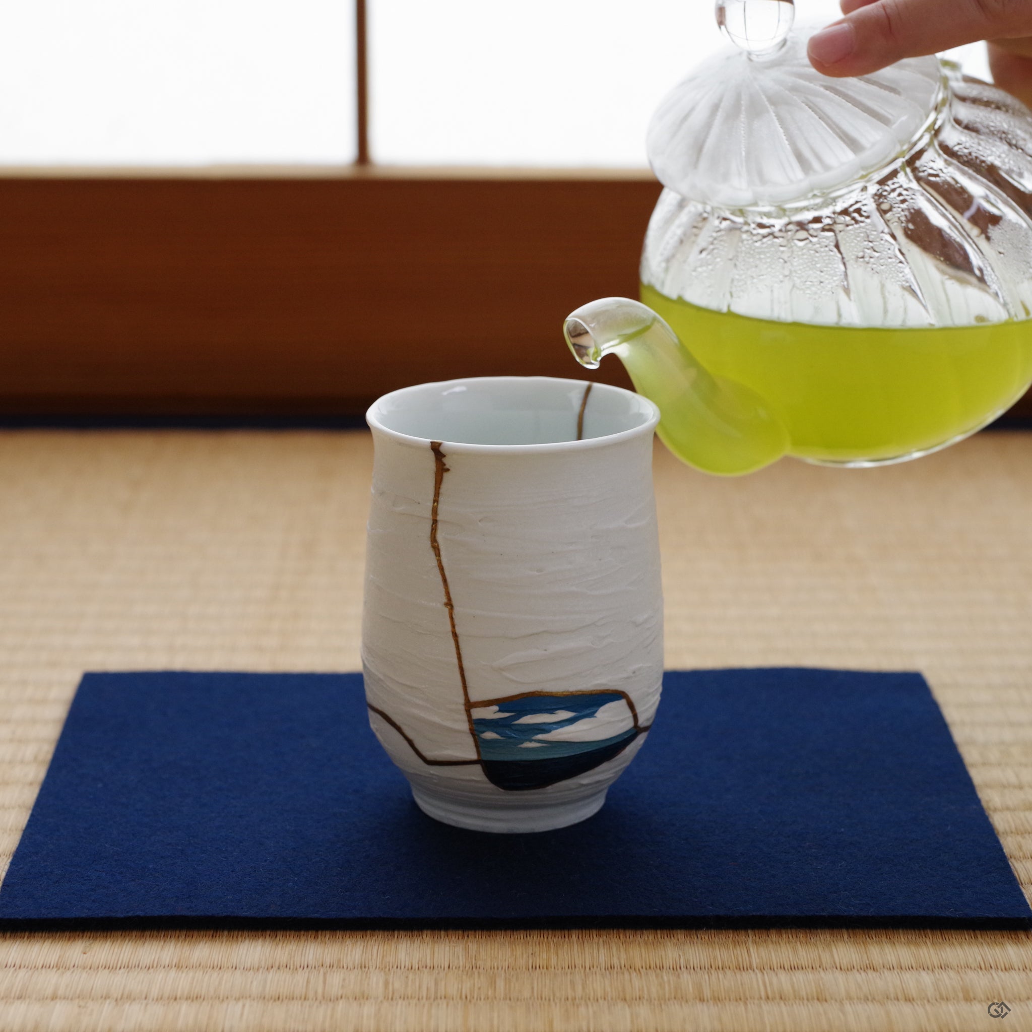 A quiet tea moment in a Japanese tatami room, as green tea is poured into a Nabeshima cup adorned with gold Kintsugi and a blue Urushi-e landscape.