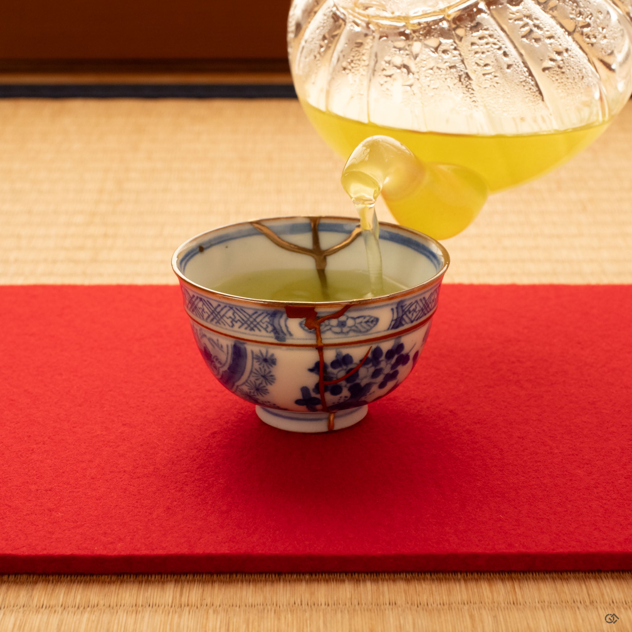 Kintsugi Ko-Imari cup receiving green tea from a glass pot, gold seams catching light in a moment of contemplation.