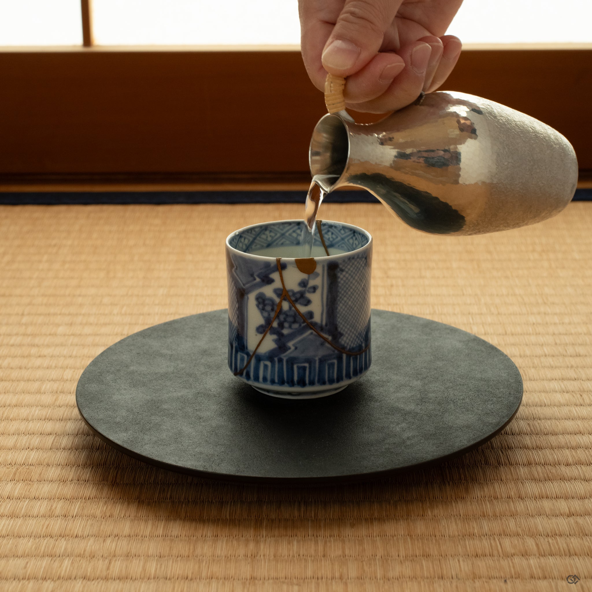 Kintsugi sake cup rests on tatami mat, illuminated by soft window light, carrying the feeling of intentional living.
