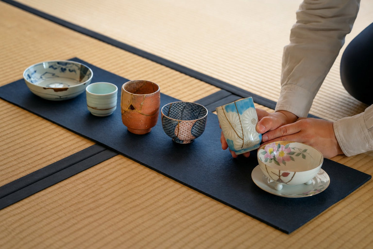 A collector choosing an authentic kintsugi gift from a row of Japanese ceramics restored with urushi and 24K gold, set on a tatami mat