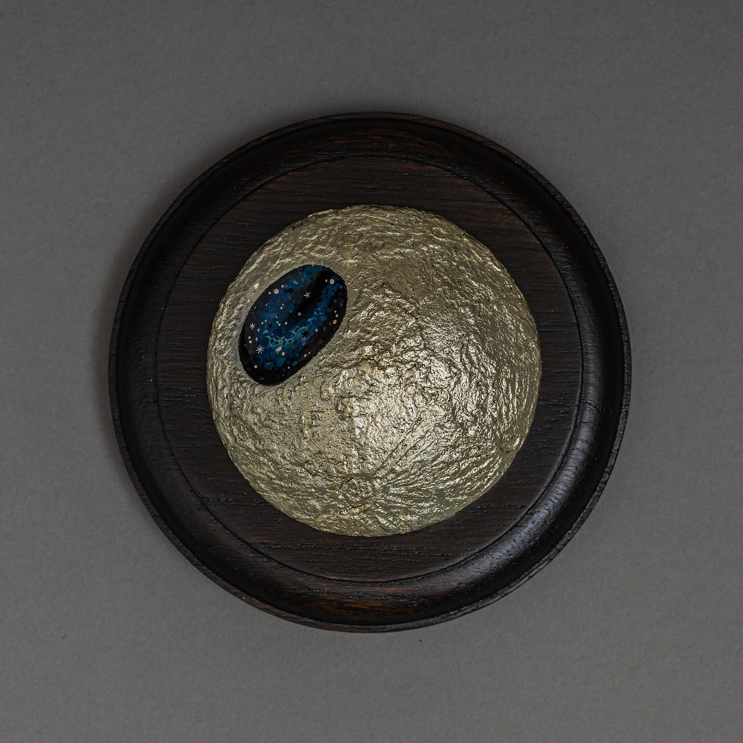 Top view of Keiko Hata's Amamitsuki (Full Moon of the Heavens, 2024) mounted on a round dark wood panel: a textured gold-leaf urushi (Japanese lacquer) moon set with an oval blue raden (mother-of-pearl) window of stars.