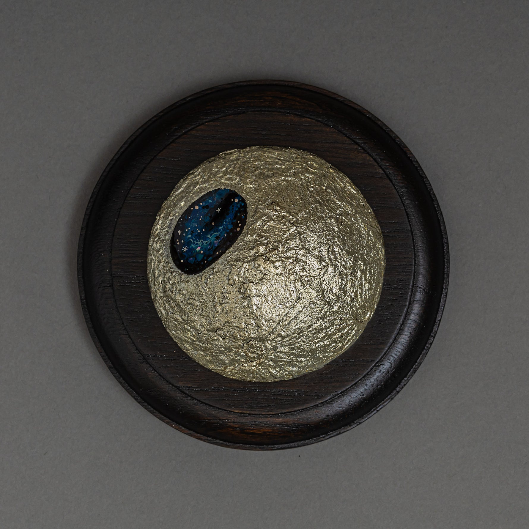 Top view of Keiko Hata's Amamitsuki (Full Moon of the Heavens, 2024) mounted on a round dark wood panel: a textured gold-leaf urushi (Japanese lacquer) moon set with an oval blue raden (mother-of-pearl) window of stars.