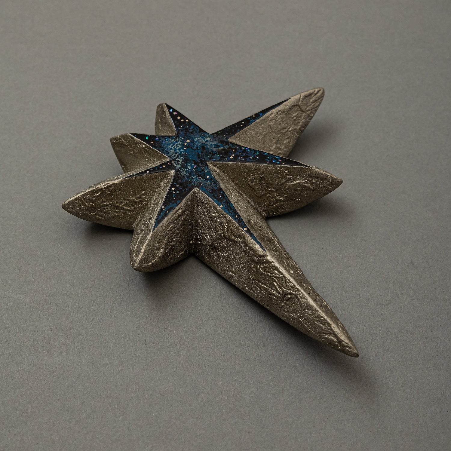 Three-quarter view of Keiko Hata's Kiraboshi / Hakugin (Glittering Stars · Silver, 2024): a multi-pointed star with textured silver tin-powder rays surrounding a blue-black urushi (Japanese lacquer) core inlaid with glittering raden (mother-of-pearl).