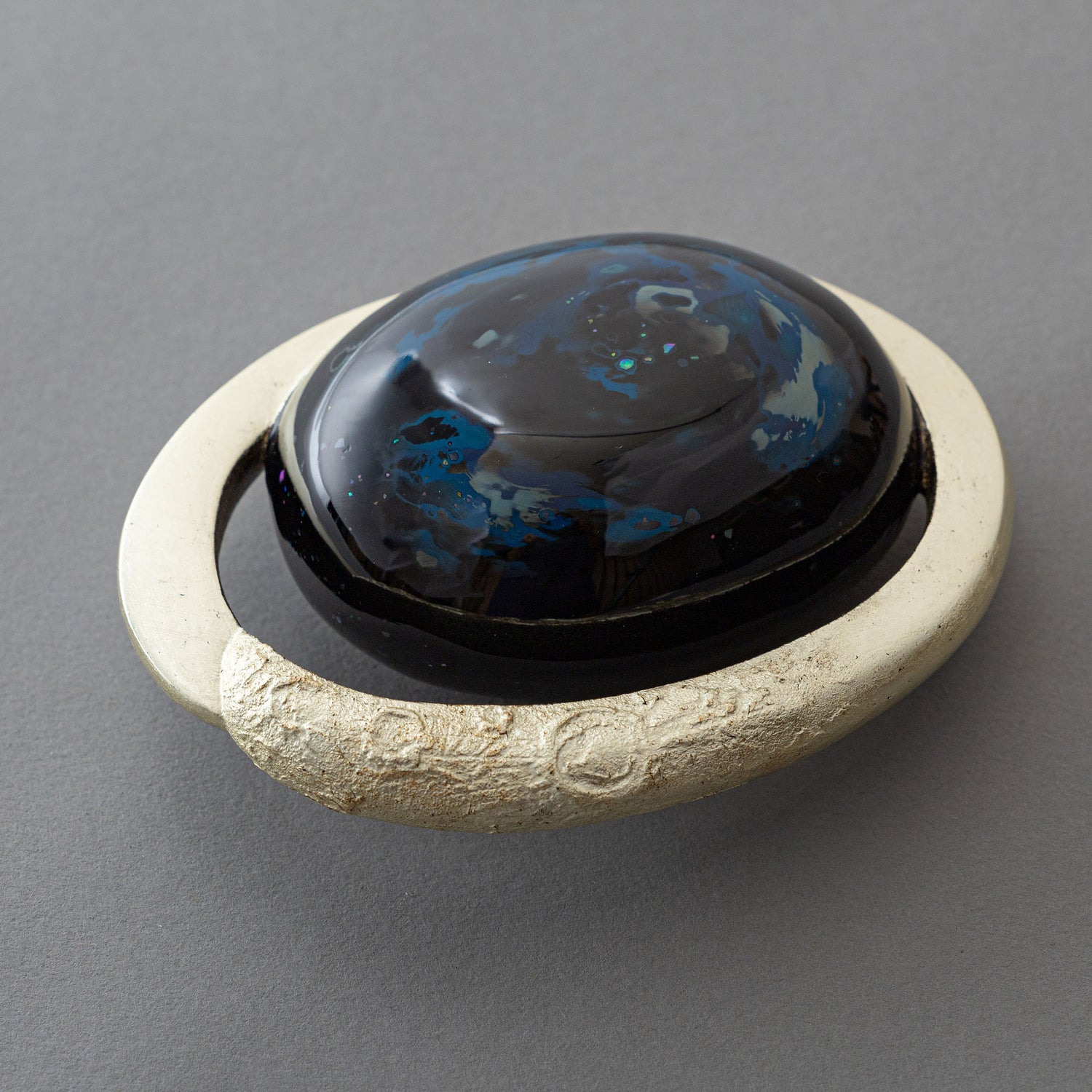 Angled view of Keiko Hata's Union of Skies (2024): a domed black urushi (Japanese lacquer) center with blue raden (mother-of-pearl) inlay, cradled in a pale gilded ring finished with water gold leaf.