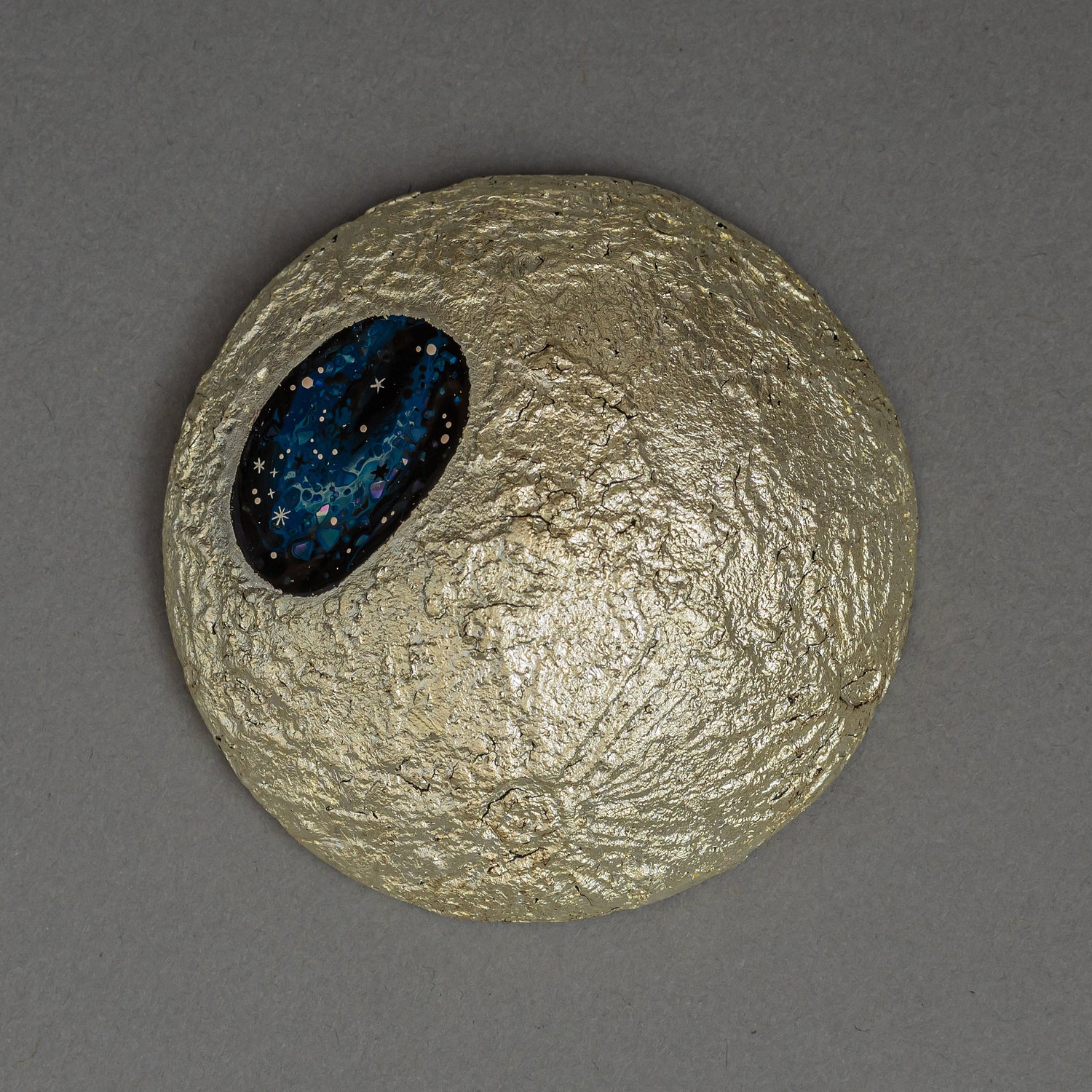 The gilded moon of Amamitsuki by Keiko Hata seen from above, its cratered gold-leaf surface holding an oval blue raden window scattered with starlike flecks.