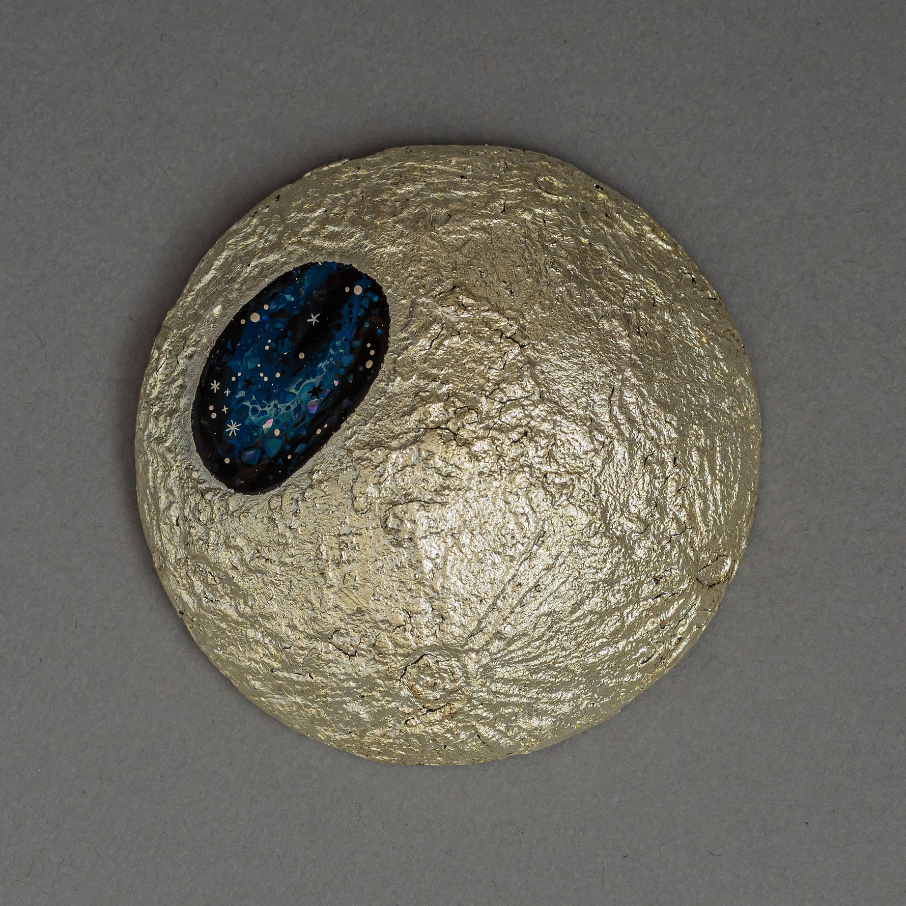 The gilded moon of Amamitsuki by Keiko Hata seen from above, its cratered gold-leaf surface holding an oval blue raden window scattered with starlike flecks.