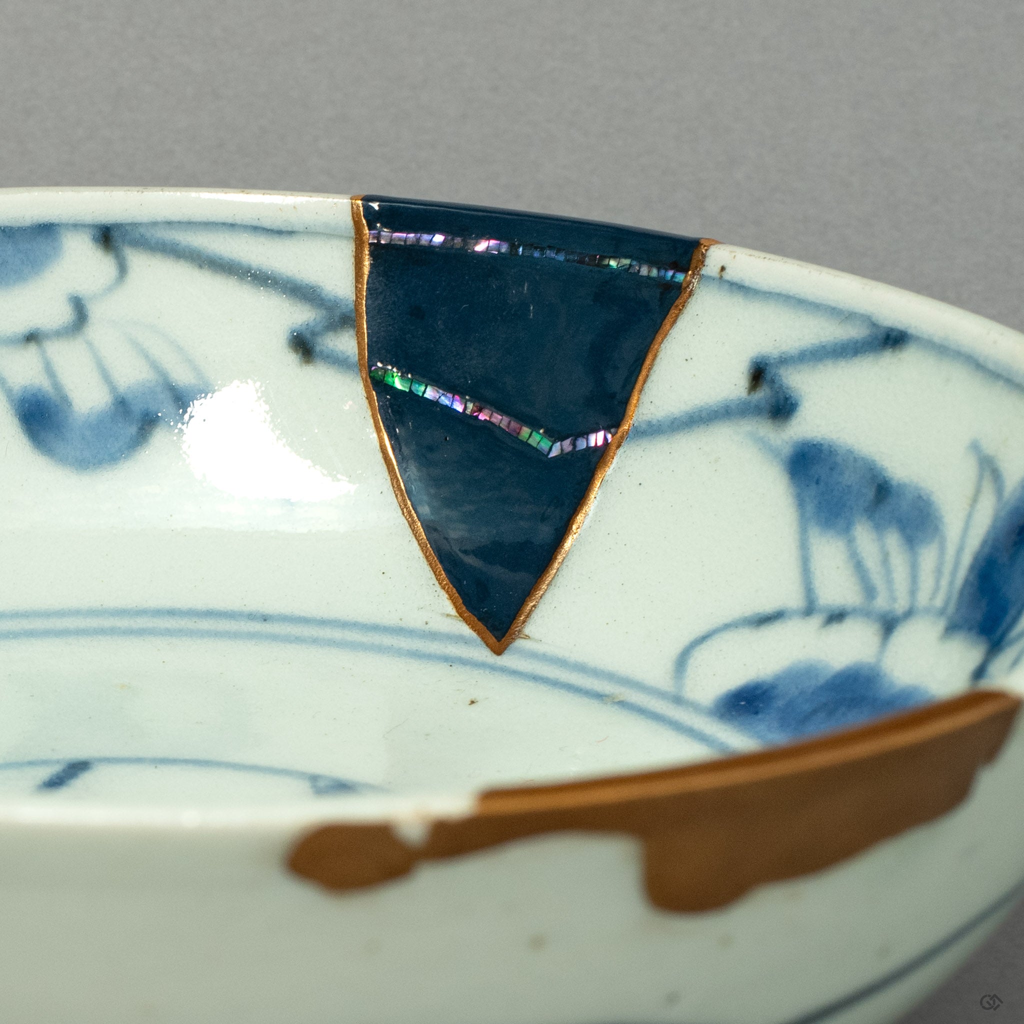 lose-up of a Ko-Imari ware kintsugi bowl showing raden (mother-of-pearl inlay) set within deep urushi lacquer, bordered by 24K gold kintsugi seams. The iridescent shell fragments catch light against the dark lacquer surface, alongside hand-painted blue and white porcelain. Restored by The Kintsugi Labo JAPAN.