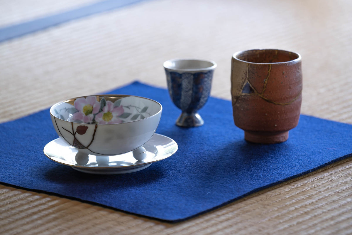 Adorned kintsugi bowl, sake cup and yunomi with decorated urushi lacquer repairs on an indigo cloth over tatami