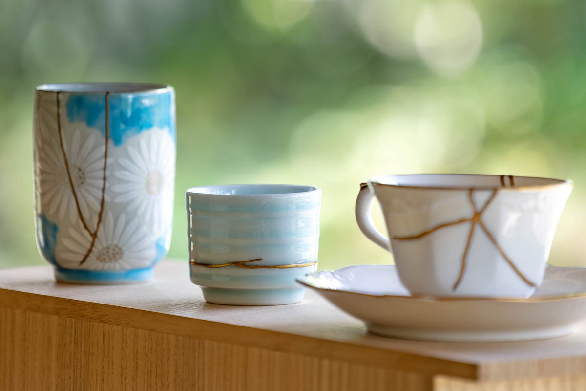 Authentic kintsugi porcelain teacups and saucer restored with natural urushi and gold, displayed on a wooden stand.