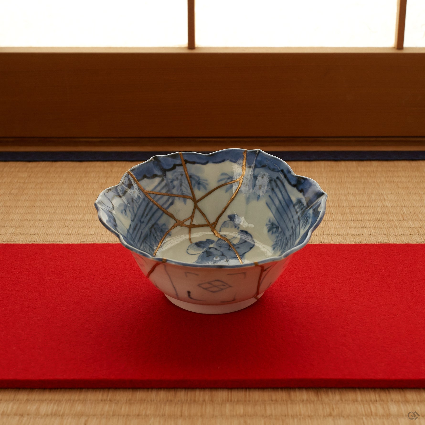 Ko-Imari blue-and-white kintsugi bowl set on a tatami mat with a red runner, its cobalt sage-and-bamboo interior and 24K gold seams shown within a quiet Japanese interior.