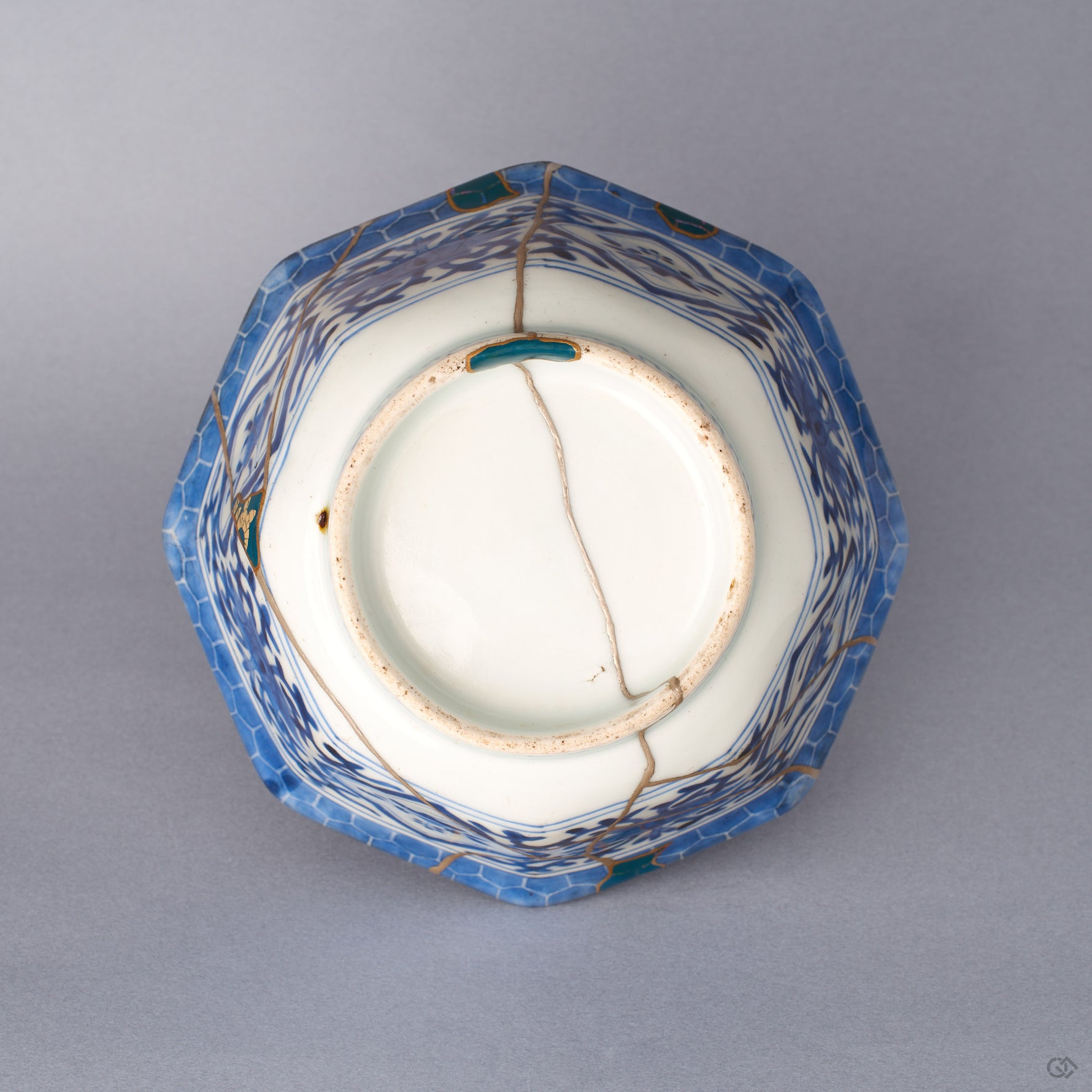 Underside of the octagonal Ko-Imari bowl, several gold-outlined green urushi fills with raden across the faceted walls and foot, silver kintsugi seams over the blue-and-white porcelain.