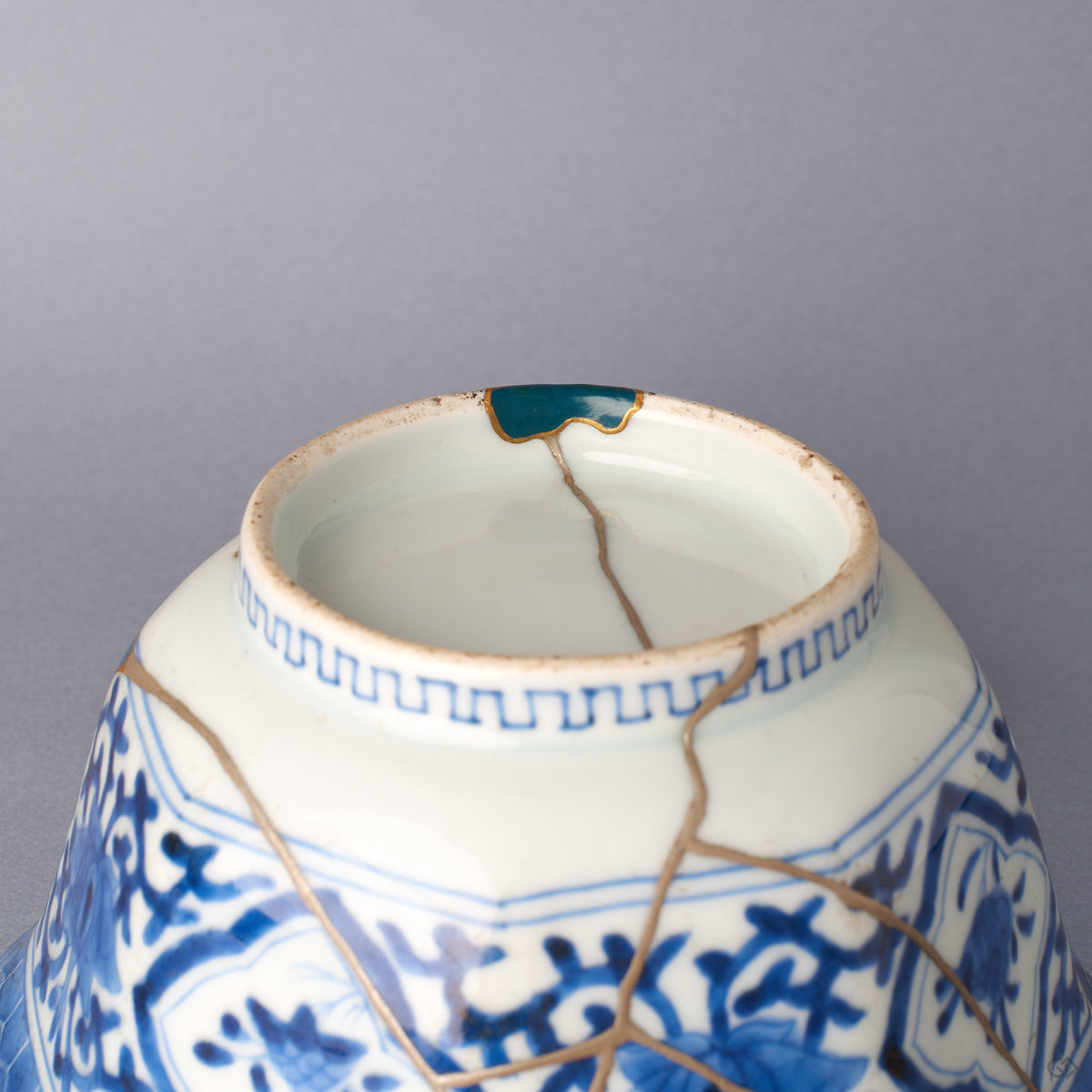 Close detail of the bowl's foot and lower body, a larger loss restored with a gold-outlined green urushi fill, the key-fret foot band and silver kintsugi seam meeting the blue-and-white glaze.