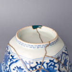 Close detail of the bowl's foot and lower body, a larger loss restored with a gold-outlined green urushi fill, the key-fret foot band and silver kintsugi seam meeting the blue-and-white glaze.