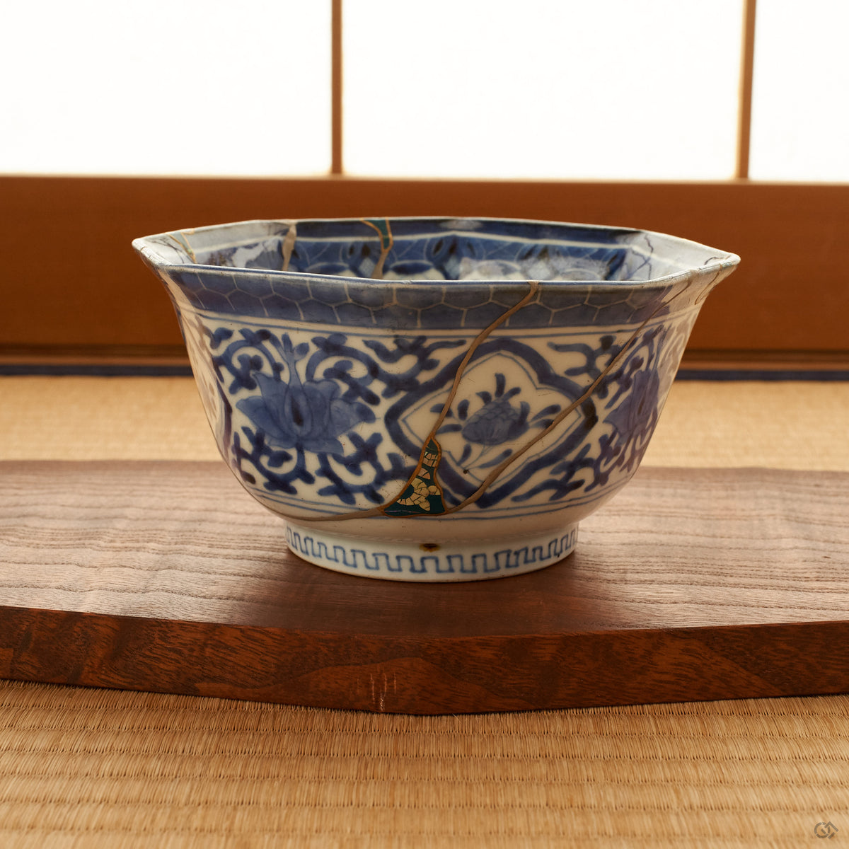 Octagonal blue-and-white Ko-Imari kintsugi bowl resting on a wooden stand on a tatami mat, a shoji screen behind it; green urushi and raden panels and 24K gold seams visible on the faceted body.