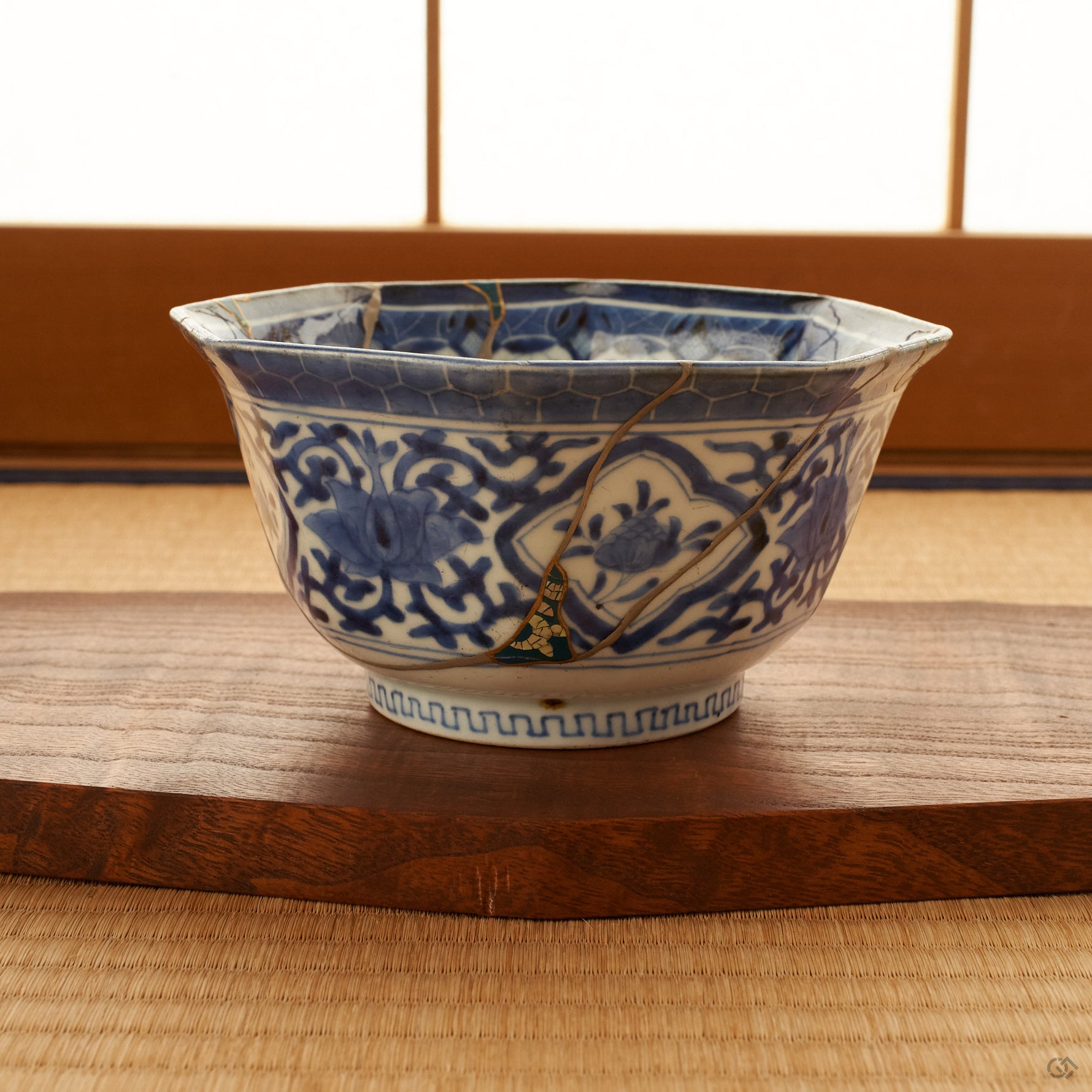 Octagonal blue-and-white Ko-Imari kintsugi bowl resting on a wooden stand on a tatami mat, a shoji screen behind it; green urushi and raden panels and 24K gold seams visible on the faceted body.