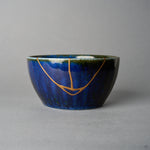 Japanese kintsugi bowl in Mino ware: a V-shaped seam of urushi and 24K gold crosses the starry night-blue kamahen glaze to the olive-green rim, front view.