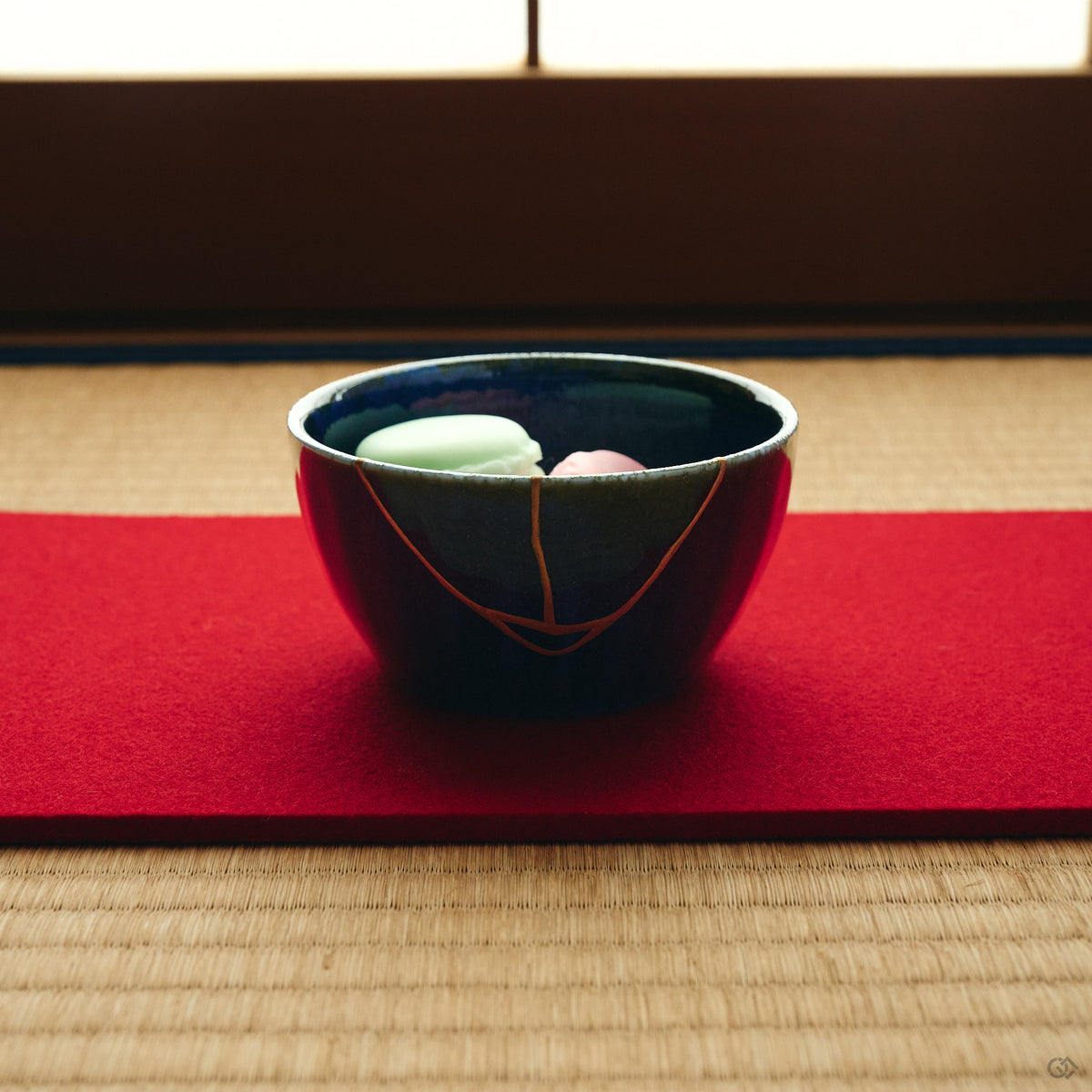 Kintsugi bowl in Mino ware holding Japanese sweets on a red felt runner, the 24K gold urushi seam climbing the night-blue wall.