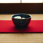 Kintsugi bowl in Mino ware holding Japanese sweets on a red felt runner, the 24K gold urushi seam climbing the night-blue wall.