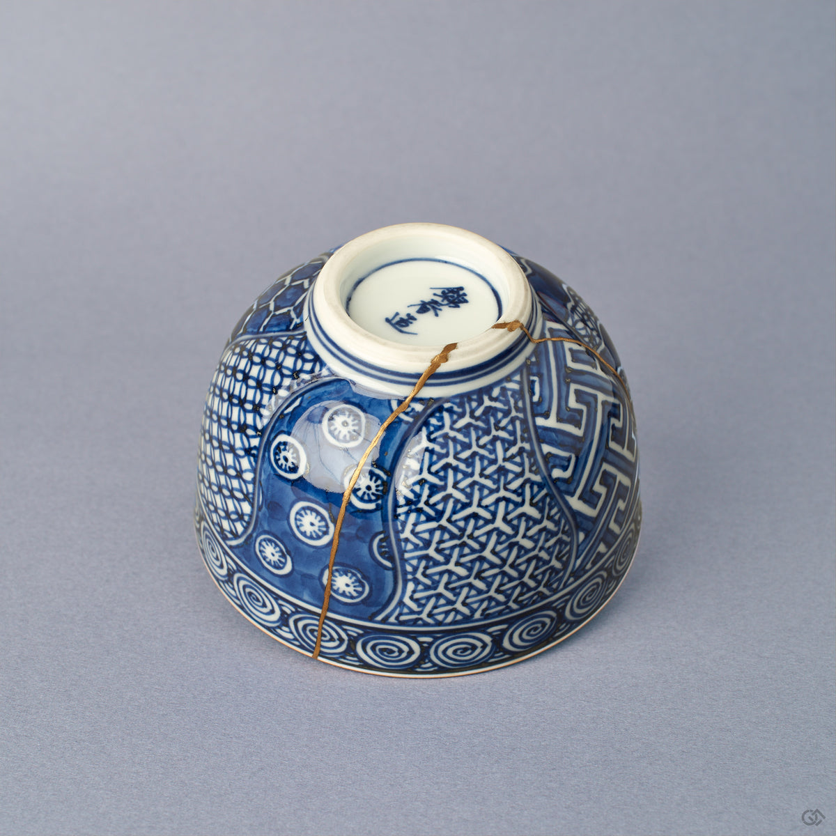 Side view of a Kyoto ware kintsugi cup, sometsuke blue with key-fret and basket-weave bands, 24K gold seam tracing the break, fitted lid.