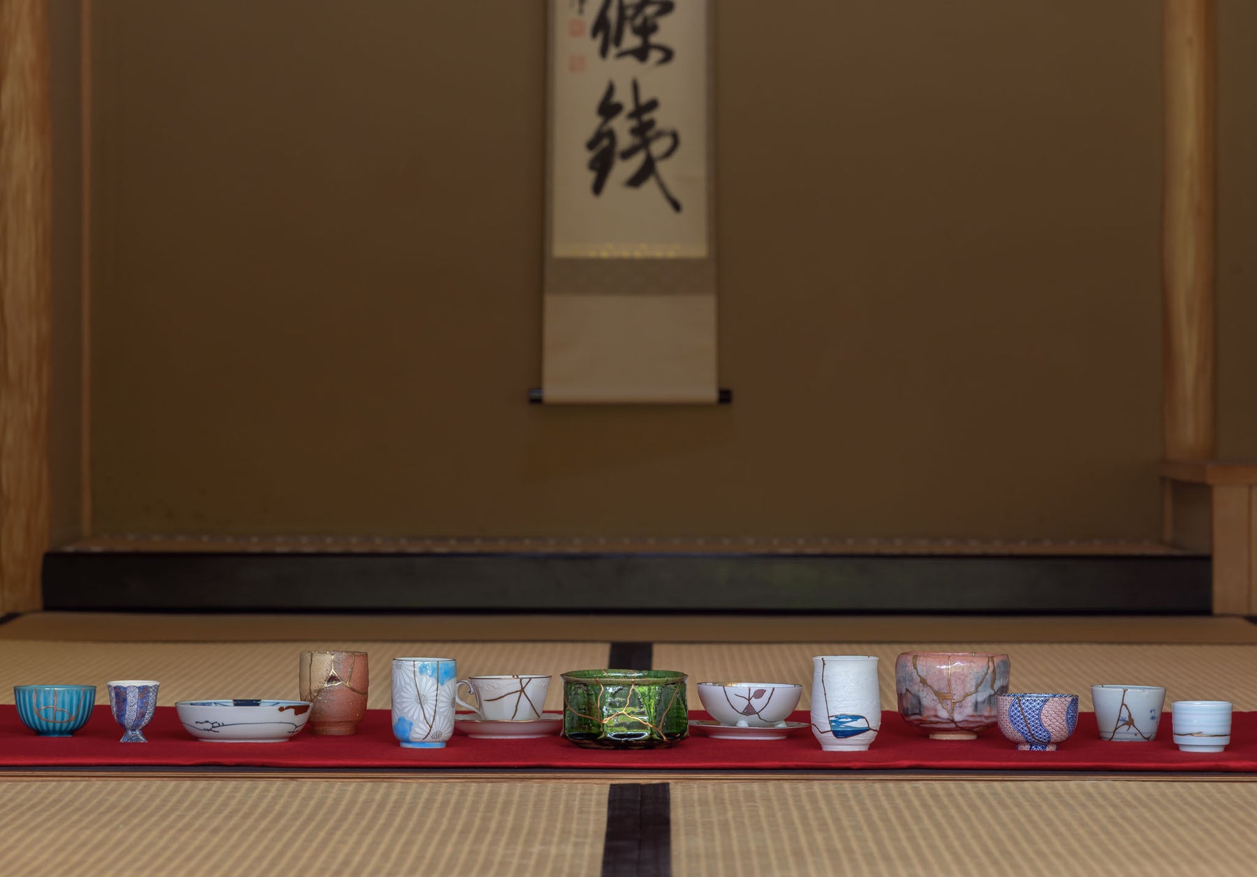 A curated collection of approximately thirteen exquisite Kintsugi masterpieces, artfully displayed in a row on a vibrant red felt cloth over traditional tatami mats. Each piece showcases the unique beauty of imperfections restored with urushi lacquer, the seams adorned with gold powder or other powdered metals, representing the timeless Japanese art of Kintsugi.