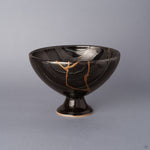 Arita ware footed bowl in glossy black porcelain, a 24K gold kintsugi seam descending and branching down the striped glaze, raised on a slender pedestal foot, shown from the front.
