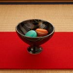 A black Arita ware footed bowl on a red runner over tatami, holding two macarons, its 24K gold kintsugi seam visible along the striped glaze, raised on a slender pedestal foot.