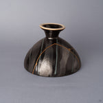 Side view of a black Arita ware footed bowl, the 24K gold kintsugi seam tracing vertically down the striped exterior glaze toward the cream-rimmed pedestal foot.