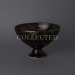 Arita ware footed bowl in glossy black porcelain with a 24K gold kintsugi seam descending the striped body, raised on a slender pedestal foot, shown from the front. Sold out.