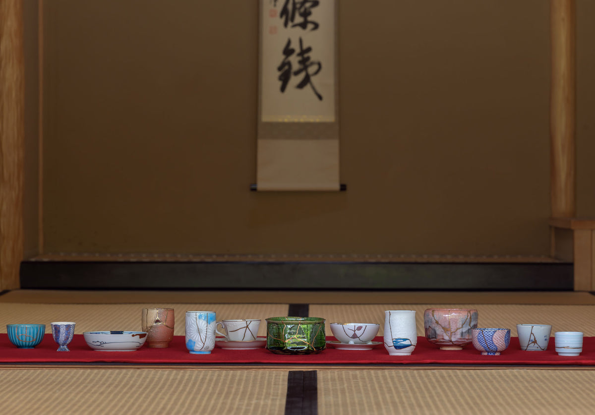 Row of authentic kintsugi cups and bowls displayed on red cloth before a hanging scroll in a Japanese tatami room