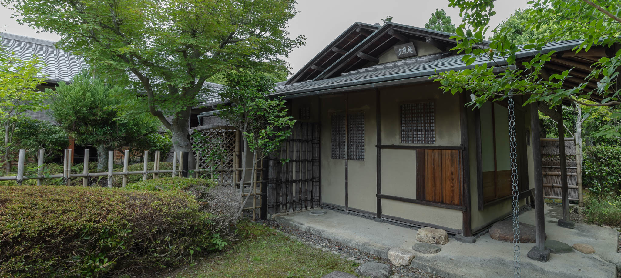 Wide-angle view of a traditional Japanese tea house (Chashitsu) exterior surrounded by a moss garden, representing the wabi-sabi aesthetic and the essence of Kintsugi and Urushi lacquerware art.