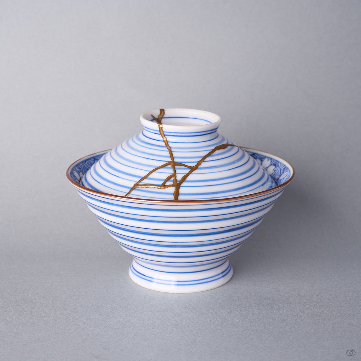 Kintsugi bowl with lid in Arita sometsuke porcelain, natural urushi and 24K gold lines descending from the knob across the blue rings of the lid, front view.