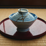 Kintsugi bowl with lid in Arita porcelain resting on a red lacquer tray over tatami, natural urushi and 24K gold lines crossing the ringed lid.