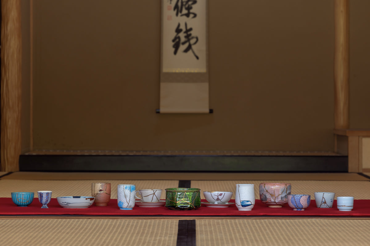 Row of authentic kintsugi cups and bowls displayed on red cloth before a hanging scroll in a Japanese tatami room