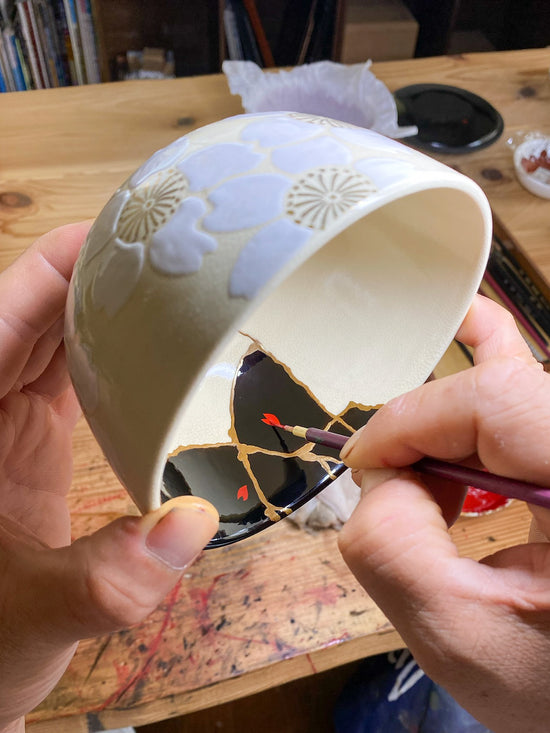 A lacquer artist painting delicate petal motifs with a maki-e brush inside a white floral matcha bowl, combining decorative urushi art with Kintsugi restoration.