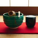 Kintsugi matcha bowl in Kyoto ware holds a bamboo whisk and scoop on a red felt mat, urushi and 24K gold seams on the emerald glaze.