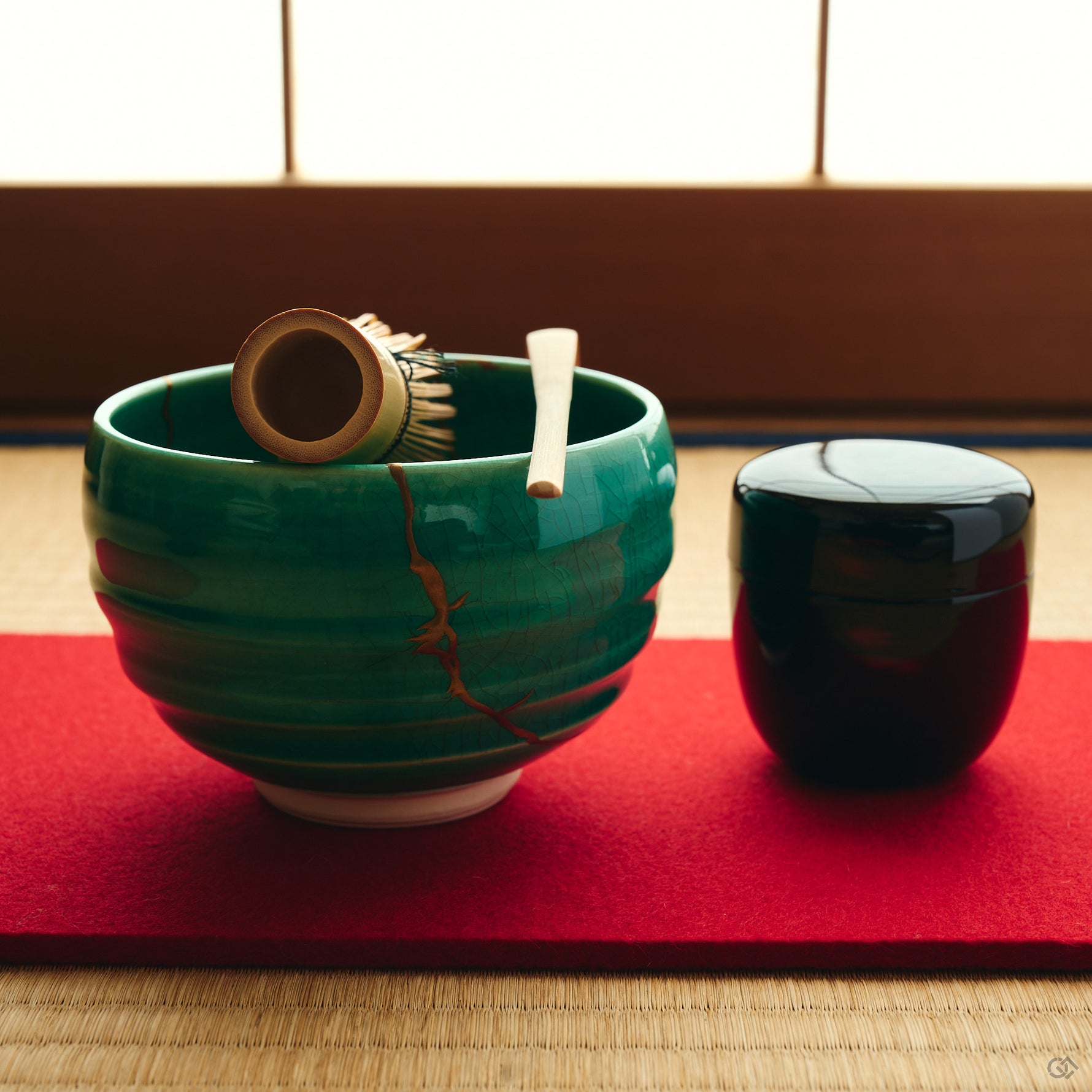 Kintsugi matcha bowl in Kyoto ware holds a bamboo whisk and scoop on a red felt mat, urushi and 24K gold seams on the emerald glaze.