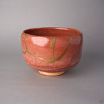 Kintsugi matcha bowl in Raku-style Kyoto ware, rejoined with natural urushi, 24K gold seams meeting in a V low on the crackled red wall, side view.