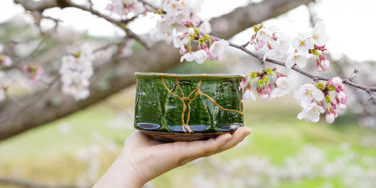 Hand holding a restored Oribe-style matcha bowl with authentic urushi Kintsugi, photographed beneath cherry blossoms—highlighting Japanese artistry and the quiet beauty of renewal.