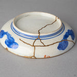 Kintsugi plate in Ko-Imari porcelain turned over, rejoined with natural urushi, branching 24K gold seams crossing the foot ring and underside, back view.