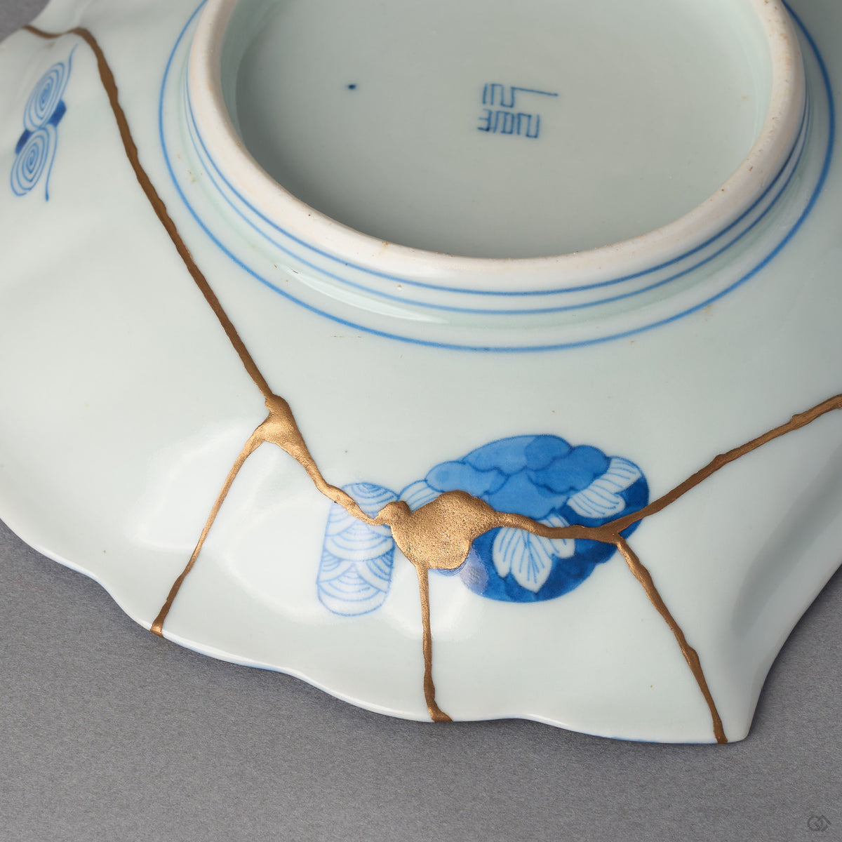 Kintsugi plate underside in Ko-Imari porcelain, rejoined with natural urushi, five 24K gold lines meeting at one point beside a blue cloud medallion, back view.