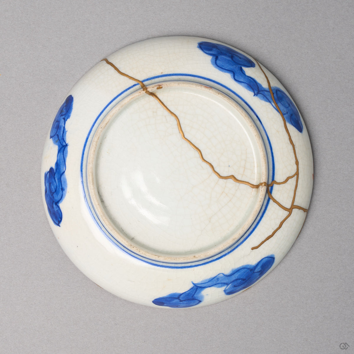 Kintsugi plate in Ko-Imari porcelain from below, rejoined with natural urushi, one 24K gold seam across the foot ring, a forked seam at the edge, bottom view.