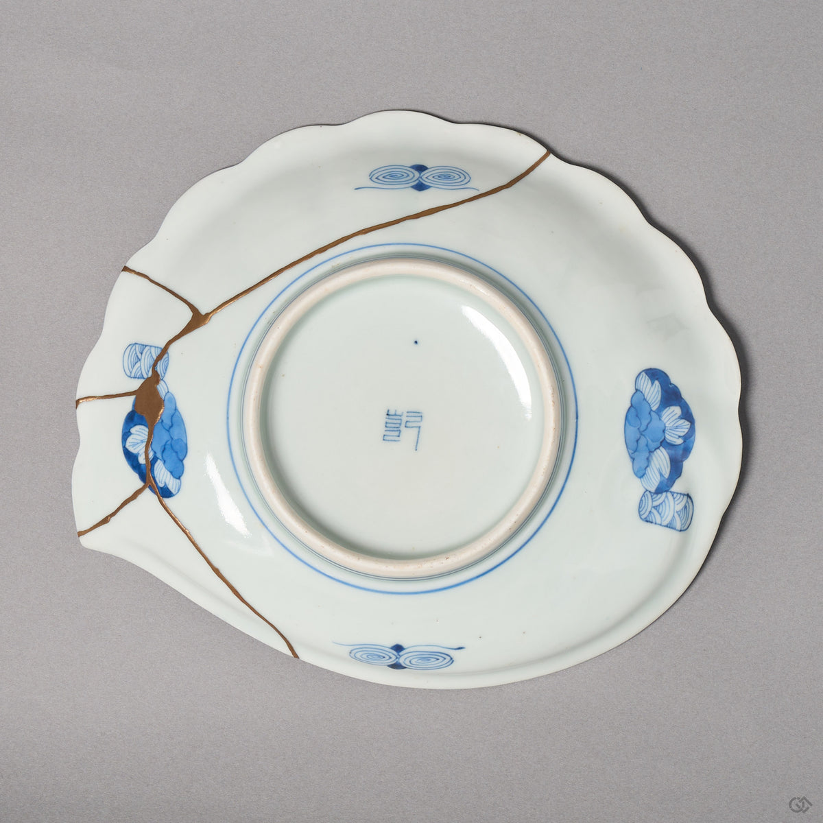 Kintsugi plate base in Ko-Imari porcelain with cloud medallions, rejoined with natural urushi, 24K gold lines meeting at one point near the edge, bottom view.