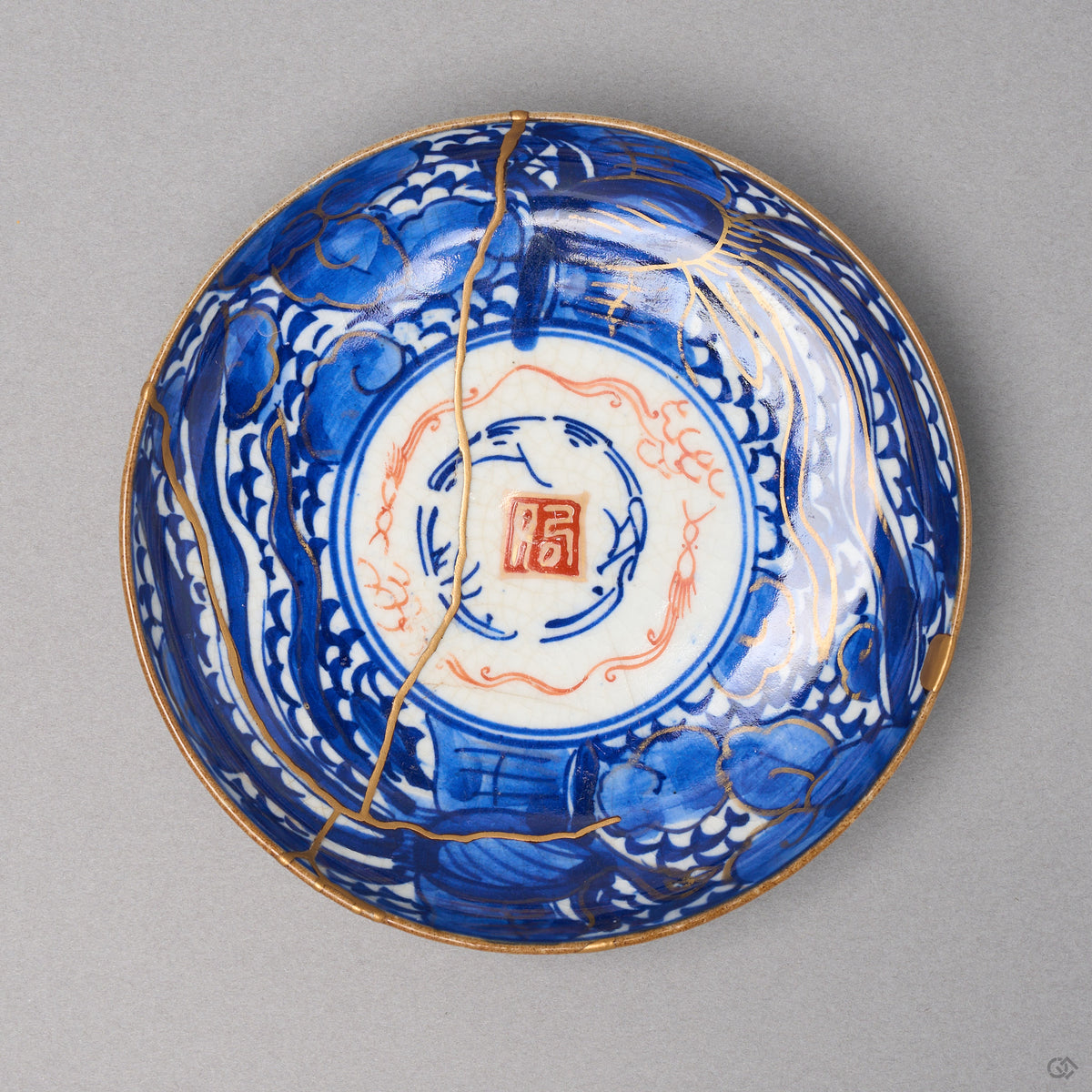 Kintsugi plate in Ko-Imari porcelain with sometsuke clouds and a red seal, rejoined with natural urushi, one 24K gold seam crossing rim to rim, front view.