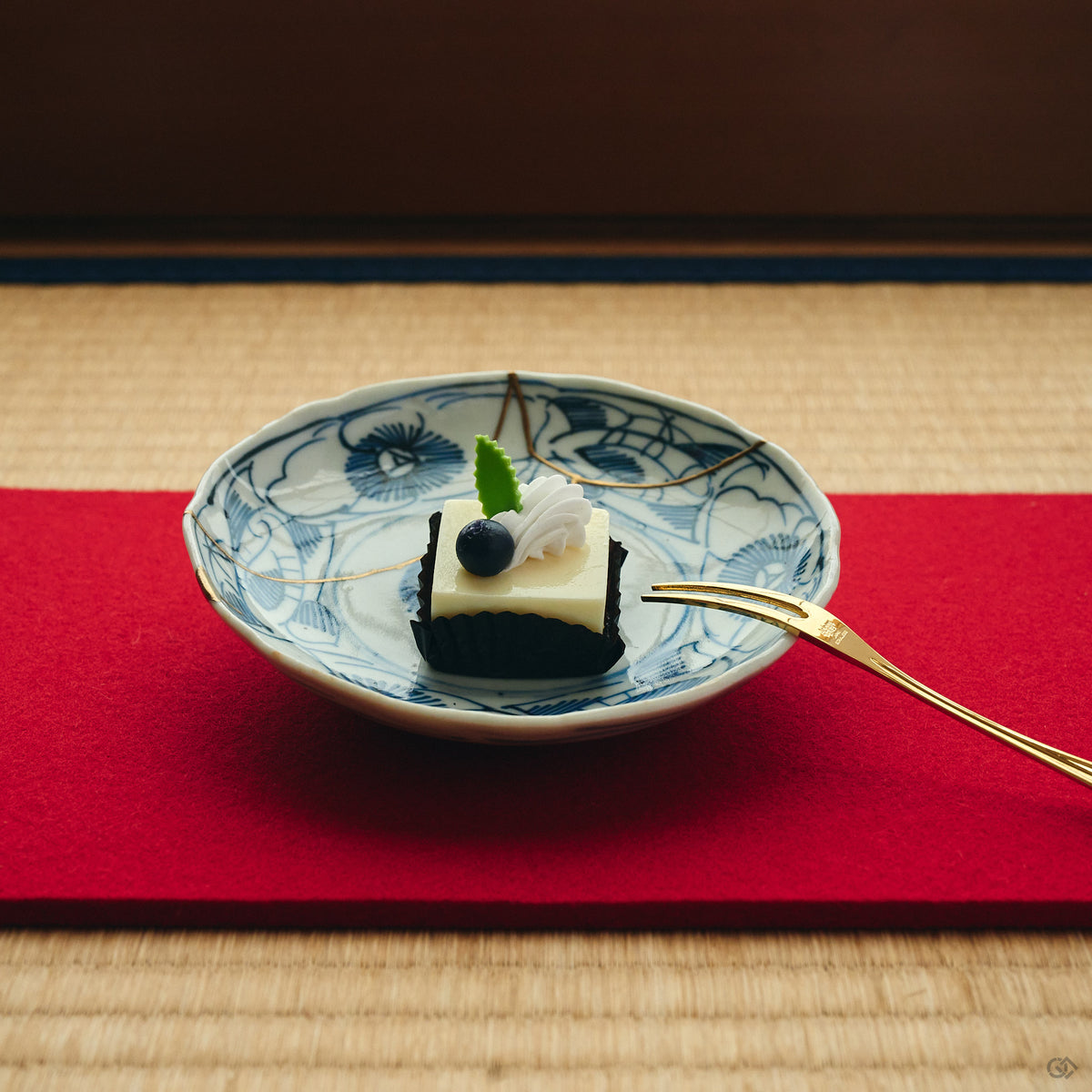 Kintsugi plate in Ko-Imari porcelain holding a small cake on a red runner over tatami, natural urushi and 24K gold seams crossing its blue camellias.