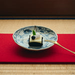 Kintsugi plate in Ko-Imari porcelain holding a small cake on a red runner over tatami, natural urushi and 24K gold seams crossing its blue camellias.