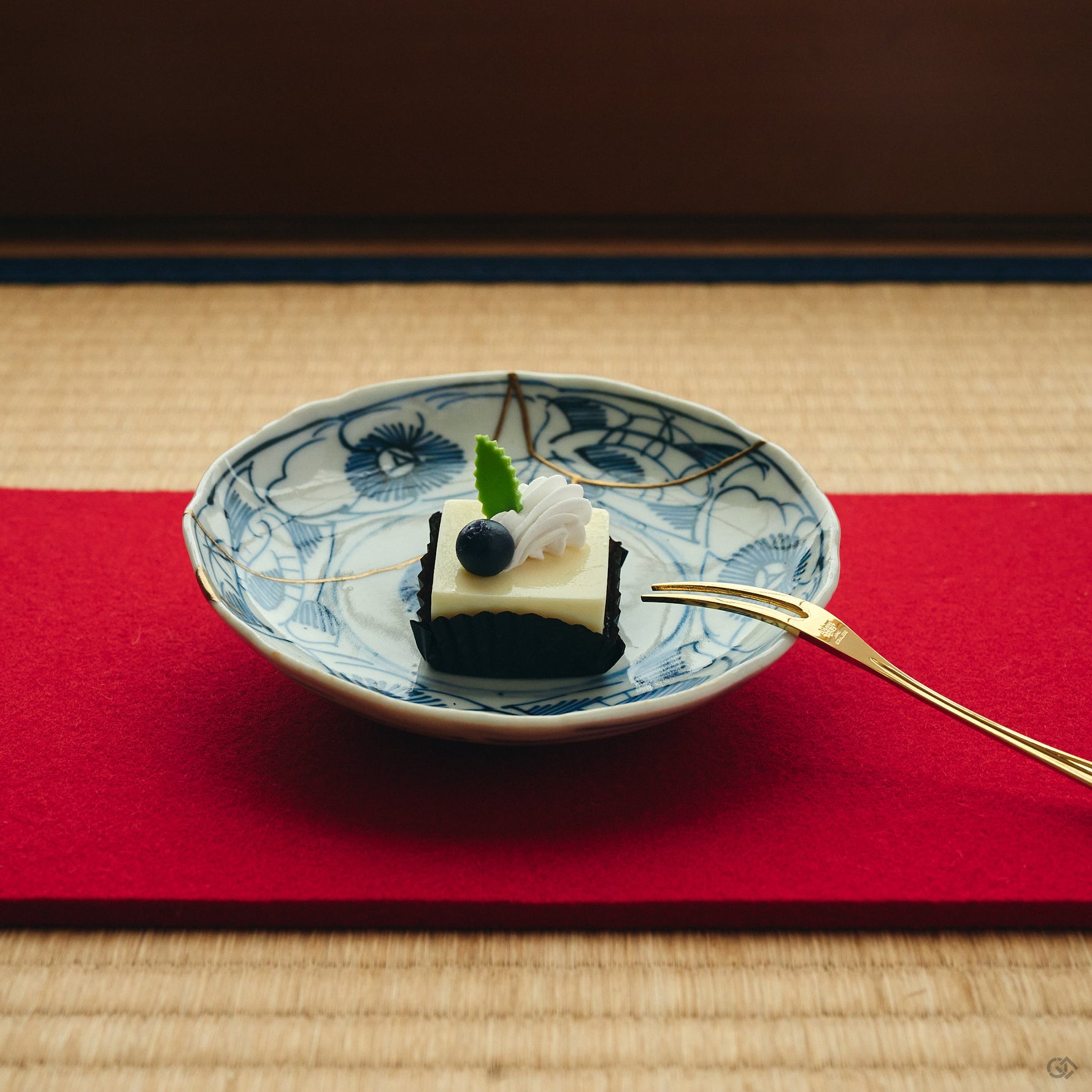 Kintsugi plate in Ko-Imari porcelain holding a small cake on a red runner over tatami, natural urushi and 24K gold seams crossing its blue camellias.