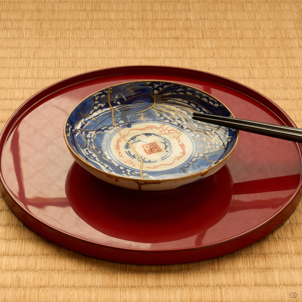 Kintsugi plate in Ko-Imari porcelain on a red lacquer tray with chopsticks, rejoined with natural urushi, a 24K gold seam crossing blue clouds, tatami setting.