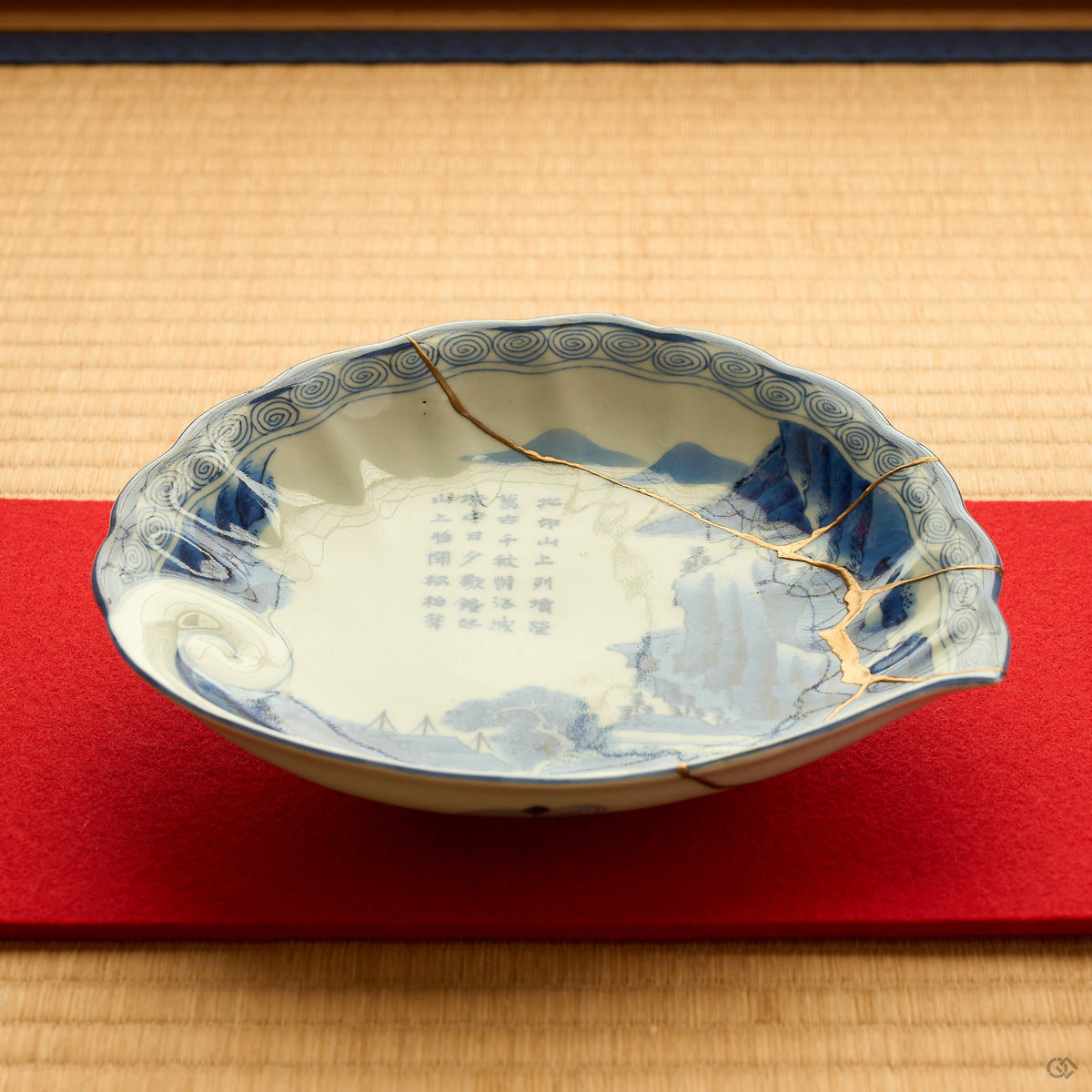 Kintsugi plate in Ko-Imari porcelain on a red felt runner over tatami, rejoined with natural urushi, the 24K gold seam descending across the painted landscape.