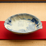Kintsugi plate in Ko-Imari porcelain on a red felt runner over tatami, rejoined with natural urushi, the 24K gold seam descending across the painted landscape.