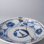 Kintsugi plate close-up in Ko-Imari porcelain, the natural urushi and 24K gold seam crossing the pine-and-plum roundel beside a gold-finished rim chip.